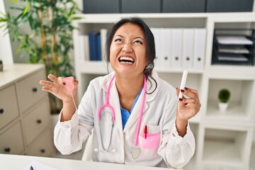 Poster - Young asian doctor woman holding menstrual cup and tampon angry and mad screaming frustrated and furious, shouting with anger looking up.