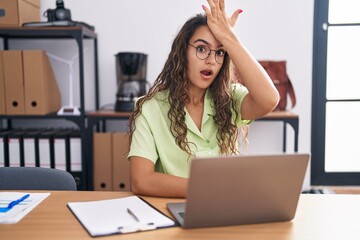 Poster - Young hispanic woman working at the office wearing glasses surprised with hand on head for mistake, remember error. forgot, bad memory concept.