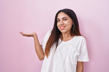 Wall Mural - Young arab woman standing over pink background smiling cheerful presenting and pointing with palm of hand looking at the camera.