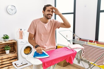 Canvas Print - Young hispanic man ironing clothes at home very happy and smiling looking far away with hand over head. searching concept.