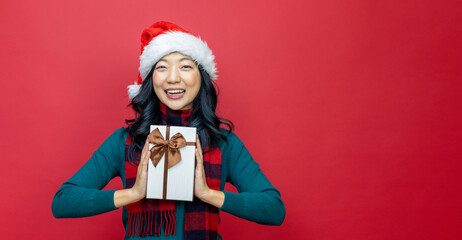 Wall Mural - Pretty smiling asian woman in warm christmas sweater and santa hat holding gift box as present with red background for season celebration concept with copy space 