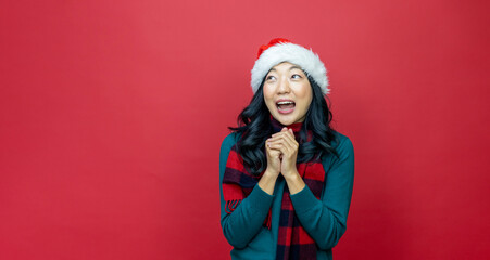 Wall Mural - Pretty smiling asian woman in warm christmas sweater and santa hat is having excitement on red background for season celebration concept with copy space 