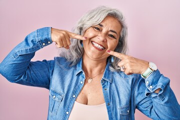 Wall Mural - Middle age woman with grey hair standing over pink background smiling cheerful showing and pointing with fingers teeth and mouth. dental health concept.