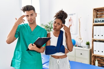 Canvas Print - Physiotherapist working with patient wearing arm on sling at rehabilitation clinic shooting and killing oneself pointing hand and fingers to head like gun, suicide gesture.