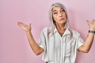 Poster - Middle age woman with grey hair standing over pink background clueless and confused expression with arms and hands raised. doubt concept.