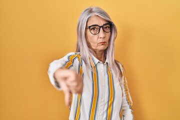 middle age woman with grey hair standing over yellow background wearing glasses looking unhappy and 