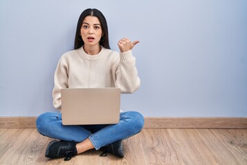 Wall Mural - Young woman using laptop sitting on the floor at home surprised pointing with hand finger to the side, open mouth amazed expression.