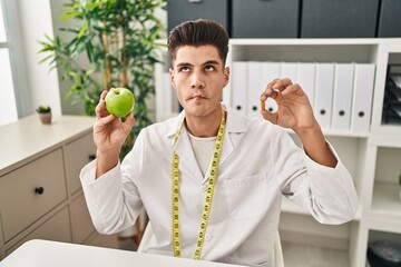 Wall Mural - Young hispanic doctor man holding pills for fat loss making fish face with mouth and squinting eyes, crazy and comical.