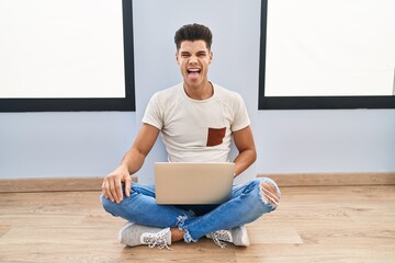 Canvas Print - Young hispanic man using laptop at home sticking tongue out happy with funny expression. emotion concept.