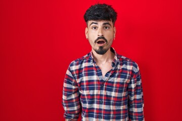 Wall Mural - Young hispanic man with beard standing over red background afraid and shocked with surprise and amazed expression, fear and excited face.