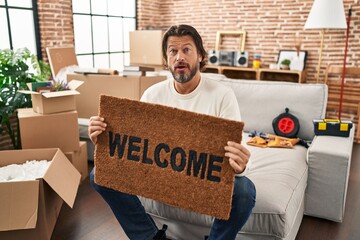 Poster - Handsome middle age man holding welcome doormat at new home in shock face, looking skeptical and sarcastic, surprised with open mouth