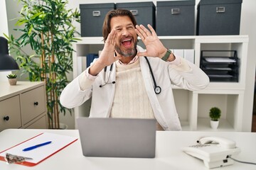 Wall Mural - Handsome middle age doctor man working at the clinic smiling cheerful playing peek a boo with hands showing face. surprised and exited