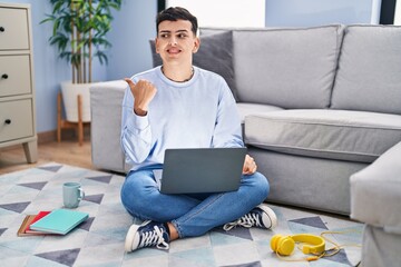 Sticker - Non binary person studying using computer laptop sitting on the floor smiling with happy face looking and pointing to the side with thumb up.