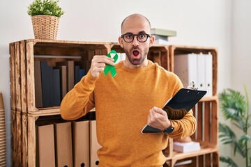 Wall Mural - Young bald man with beard holding support green ribbon afraid and shocked with surprise and amazed expression, fear and excited face.