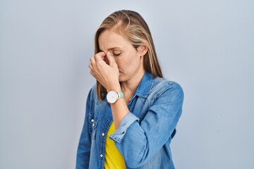 Poster - Young blonde woman standing over blue background tired rubbing nose and eyes feeling fatigue and headache. stress and frustration concept.