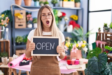 Wall Mural - Young blonde woman working at florist holding open sign in shock face, looking skeptical and sarcastic, surprised with open mouth