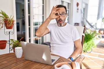 Wall Mural - Middle age man using computer laptop at home doing ok gesture shocked with surprised face, eye looking through fingers. unbelieving expression.