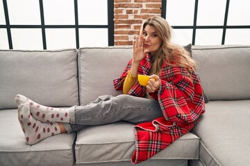 Canvas Print - Young woman sitting on the sofa drinking a coffee at home hand on mouth telling secret rumor, whispering malicious talk conversation