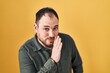 © Krakenimages.com - Plus size hispanic man with beard standing over yellow background hand on mouth telling secret rumor, whispering malicious talk conversation