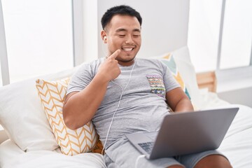 Poster - Chinese young man using laptop sitting on the bed smiling happy pointing with hand and finger