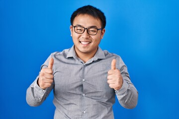 Sticker - Young chinese man standing over blue background success sign doing positive gesture with hand, thumbs up smiling and happy. cheerful expression and winner gesture.