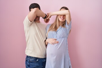 Young couple expecting a baby standing over pink background smiling cheerful playing peek a boo with hands showing face. surprised and exited