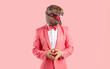 Portrait of a funny bizarre man wearing a goofy rubber dinosaur mask and a trendy pink suit with a bowtie standing isolated on a pink colour background