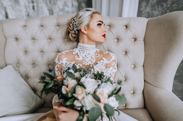 A beautiful young bride model blonde in a long lace dress sits on a sofa in the interior with a bouquet in her hands. Wedding photography, portrait.