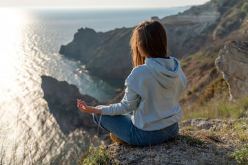 Wall Mural - Woman tourist enjoying the sunset over the sea mountain landscape. Sits outdoors on a rock above the sea. She is wearing jeans and a blue hoodie. Healthy lifestyle, harmony and meditation