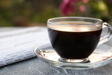 Wall Mural - Glass cup with coffee and newspaper on grey table outdoors, closeup. Morning ritual