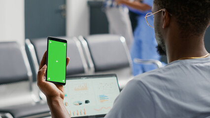 Male patient using greenscreen template on smartphone while he waits in facility waiting area. Young man looking at isolated mockup display and chroma key, blank copyspace on mobile phone screen.
