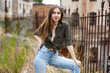 Sensual young woman with long hair wearing jeans and unbuttoned blouse knotted under her breasts posing against abandoned old house, standing near metal fence