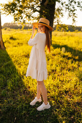 Wall Mural - Vertical portrait of gorgeous redhead young woman in straw hat and white dress standing posing on beautiful meadow of green grass looking at camera, on background of warm sunlight at summer day.