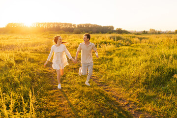Wall Mural - happy young couple in love running looking at each other in green filed during golden sunset on summ