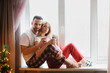 © natalialeb - Cute European couple with mugs with hot cocoa in red pajamas on window in living room. Cozy good Christmas morning. Looking at camera.