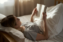 Caucasian Woman Lying Down In Bed And Reading Book At The Morning