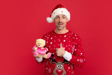 Portrait Of Christmas Santa Man Hold Fluffy Toy, Gift For Baby Kids On Red Studio Background.