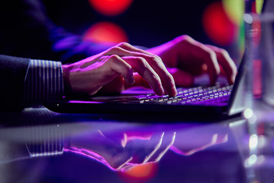 Cropped image of male hands of business man typing on laptop keyboard at evening time. Cool colored neon hue lights in office space.