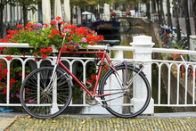 Bike Along White Metal Railing On Stone Bridge With Colourful Flowers In Box And Canal In The Background Below; Delft, South Holland, Netherlands