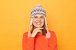 © sementsova321 - Portrait of Caucasian young adult woman wearing orange jumper and knitted earflap hat standing isolated over yellow background, keeps hand under chin, looking at camera with charming smile.
