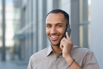 Close-up photo of a smiling man outside an office building happily talking on the phone, a businessman in a casual shirt hispanic looking at the camera portrait.