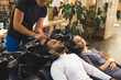 © PoppyPix - Happy indian male client having his hair washed with shampoo by male hairdresser next to blonde female customer looking into the camera in a professional hair salon. High quality photo