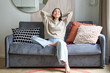 Portrait of carefree asian woman enjoying day-off, sitting on sofa and smiling pleased, relaxing in house on weekends
