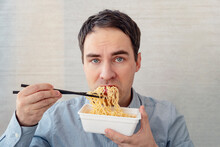 Young Man In A Blue Shirt Is Eating Noodles From A Box With A Dissatisfied Face. Lunch At The Office. Tasteless Junk Food