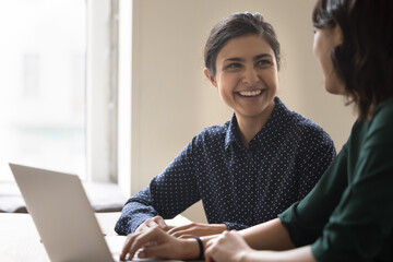 Happy young Indian employee woman talking to colleague, smiling, laughing, discussing career success, hiring, job achieve. Mentor and intern enjoying training for tasks