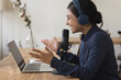 © fizkes - Happy pretty Indian blogger girl in blue wireless headphones speaking at laptop and professional microphone, talking on making video call, recording podcast, gesturing, smiling, laughing
