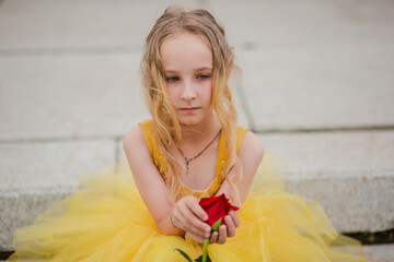 Wall Mural - A little girl with blond hair in a lush yellow dress walks in the park with a red rose. Princess image. Beautiful child in historical dress