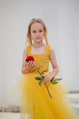 Wall Mural - A little girl with blond hair in a lush yellow dress walks in the park with a red rose. Princess image. Beautiful child in historical dress