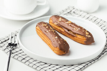Wall Mural - Eclairs with custard and chocolate icing on a white plate. Eclairs with cream filling covered with melted milk chocolate. A cup of tea with traditional French pastry in the morning. French breakfast