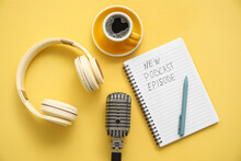 Notebook with text NEW PODCAST EPISODE, microphone, coffee cup and headphones on yellow background